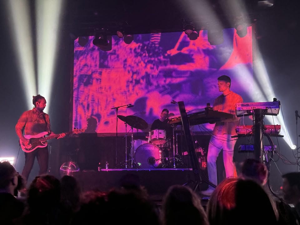 3 people on stage; one wielding a guitar, one behind a drumset, and another behind a keyboard. They are illuminated by purple and indigo lights