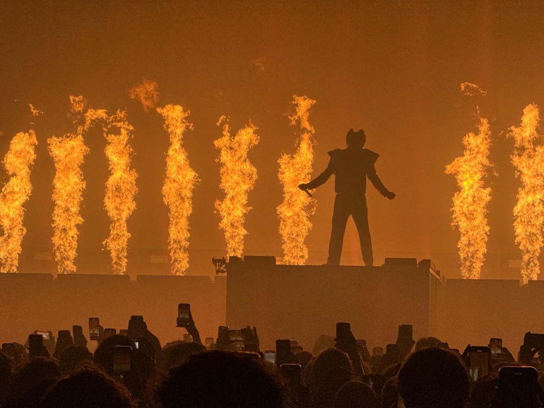 Tyler, The Creator with his arms stretched out looking commanding over the crowd visible in the foreground. He's backlit by several 3m high flames