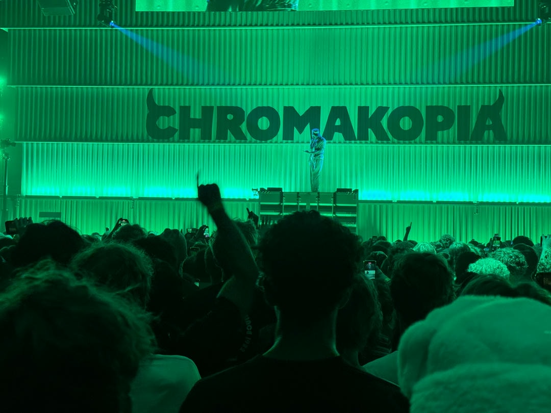 Tyler, The Creator visible on stage, with a crowd in the foreground looking at him. Behind him is a massive logotype spelling "CHROMAKOPIA" on big shipping containers