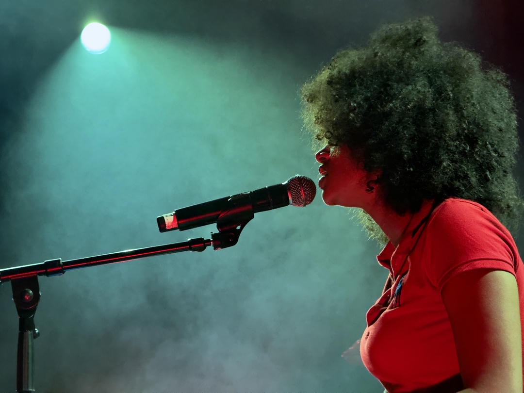 A close-up of Laila sitting at a piano, singing into a microphone. A light is also shown that's acting as a backlight, giving the photo a professional look