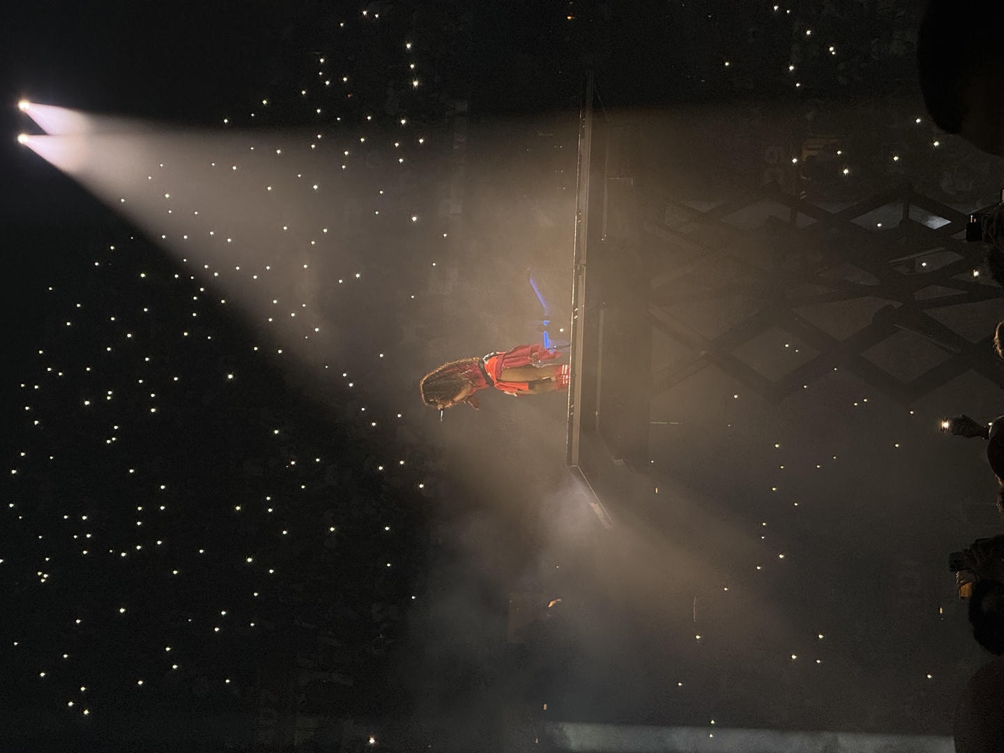 SZA lifted up on a scissor lift performing in a red outfit. She is illuminated by 2 spotlights above her.