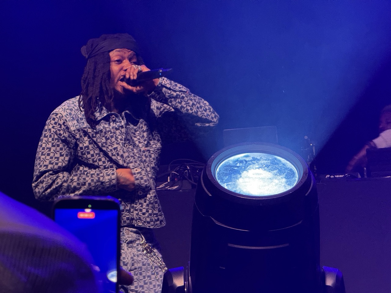JID performing live on stage. He's holding the mic up to his mouth. A phone can be seen recording the show in the foreground.
