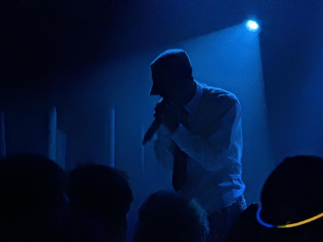 SWIM performing live on stage. Backlit by a blue spotlight, he's holding the microphone close to his face.