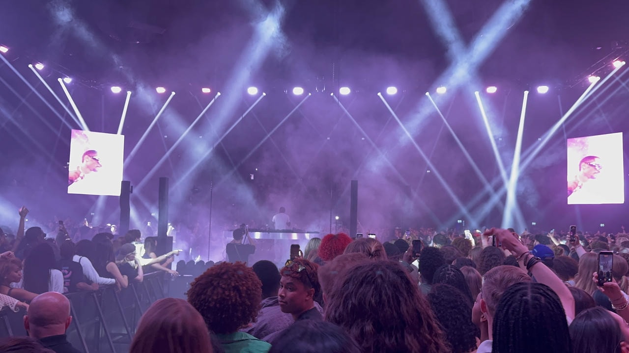 A wide-angle shot of Kaytranada behind a DJ booth in the distance. The background is filled with indigo smoke, and the foreground has a bunch of people watching his performance.