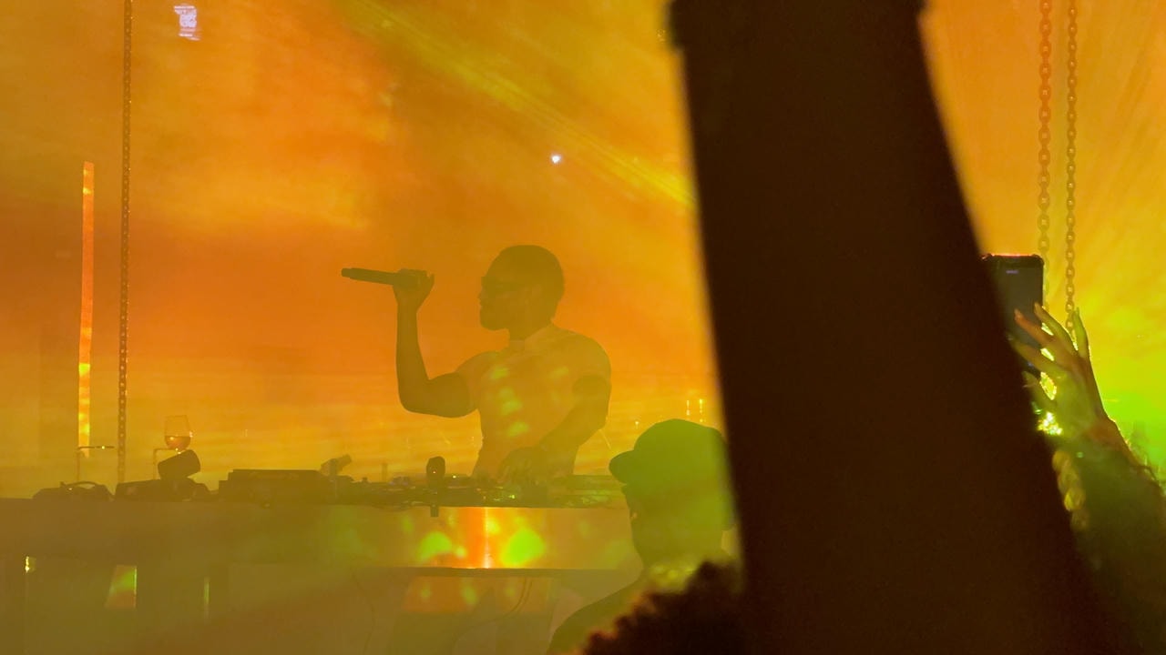 A close-up of Kaytranada DJ-ing with a yellow and orange background. He's holding the mic, grooving out to his own music.