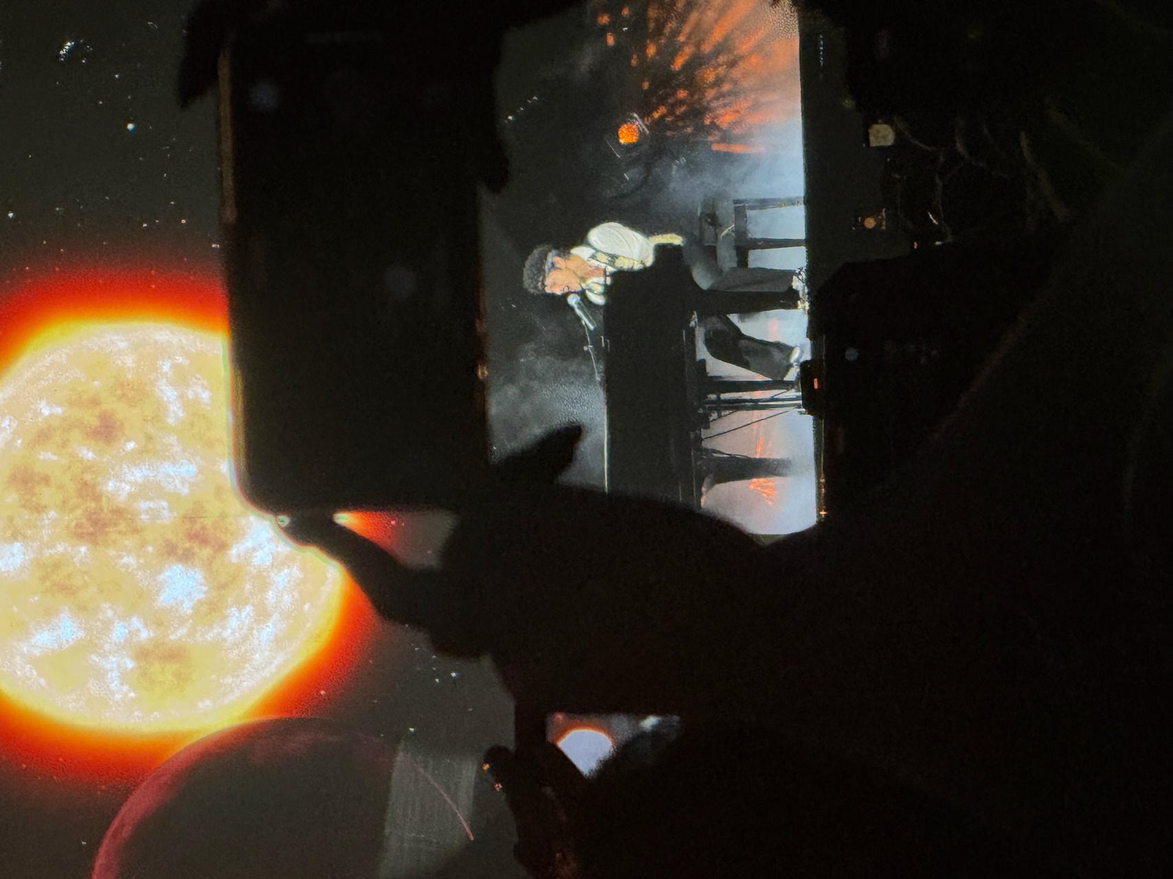Bruno Mars, sitting by a piano singing into a microphone. The background shows the sun projected on a large LED screen behind him, the foreground has people partially blocking the view with their phones.