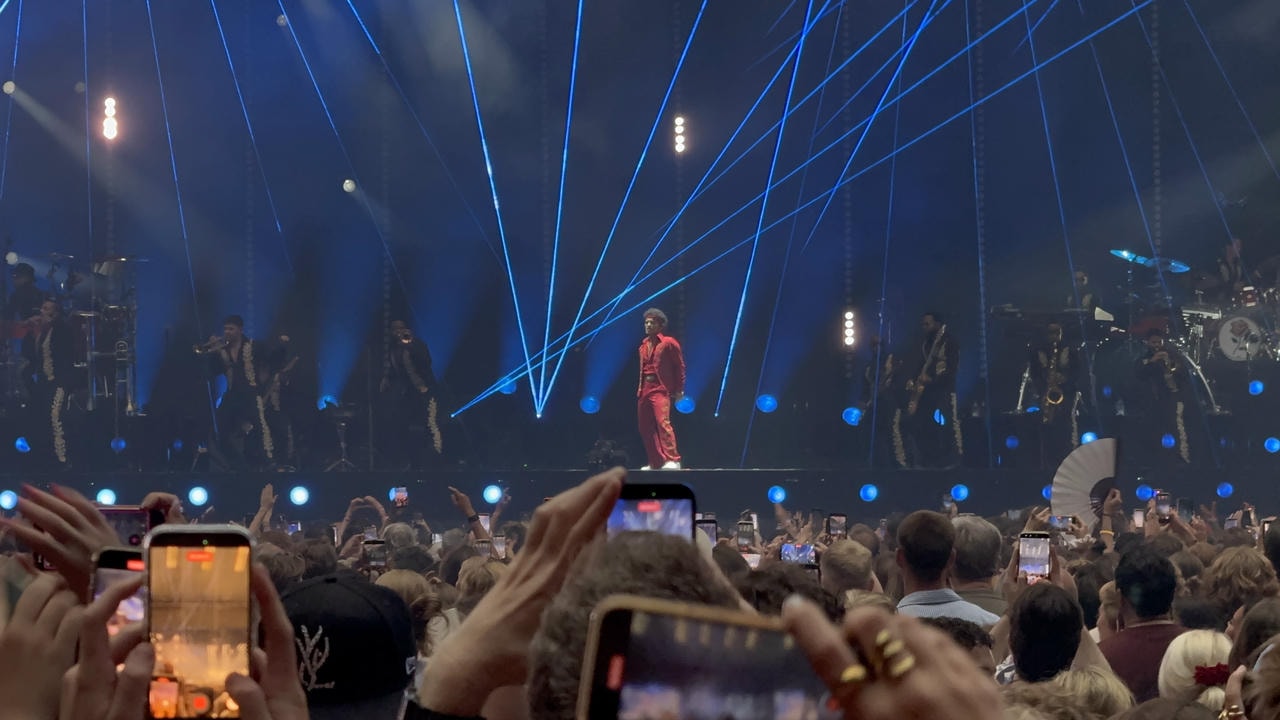 Bruno Mars in a red suit performing on stage surrounded by his band The Hooligans. There are blue lasers in the background pointing up.