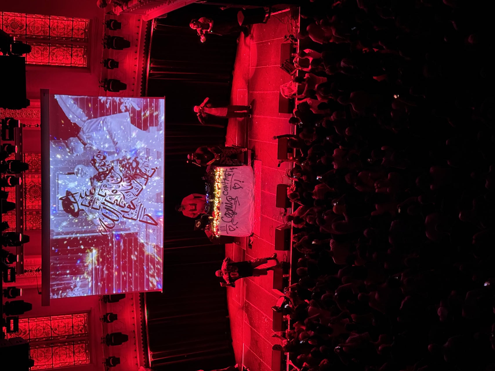 Yasiin Bey performing live on stage. On the side, Talib Kweli and The Alchemist are looking on.