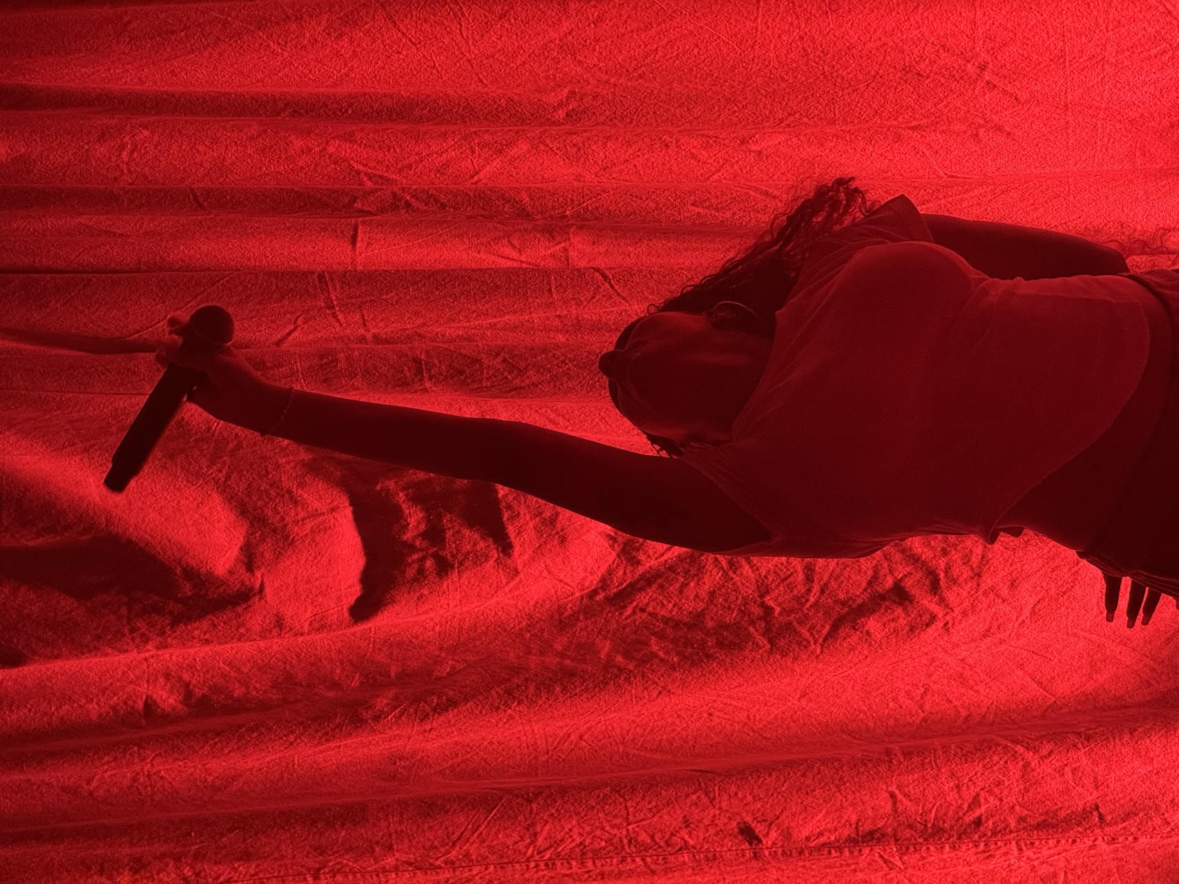 Samara Cyn posing in front of a red-lit curtain. She's reaching up with her mic, looking directly upwards.
