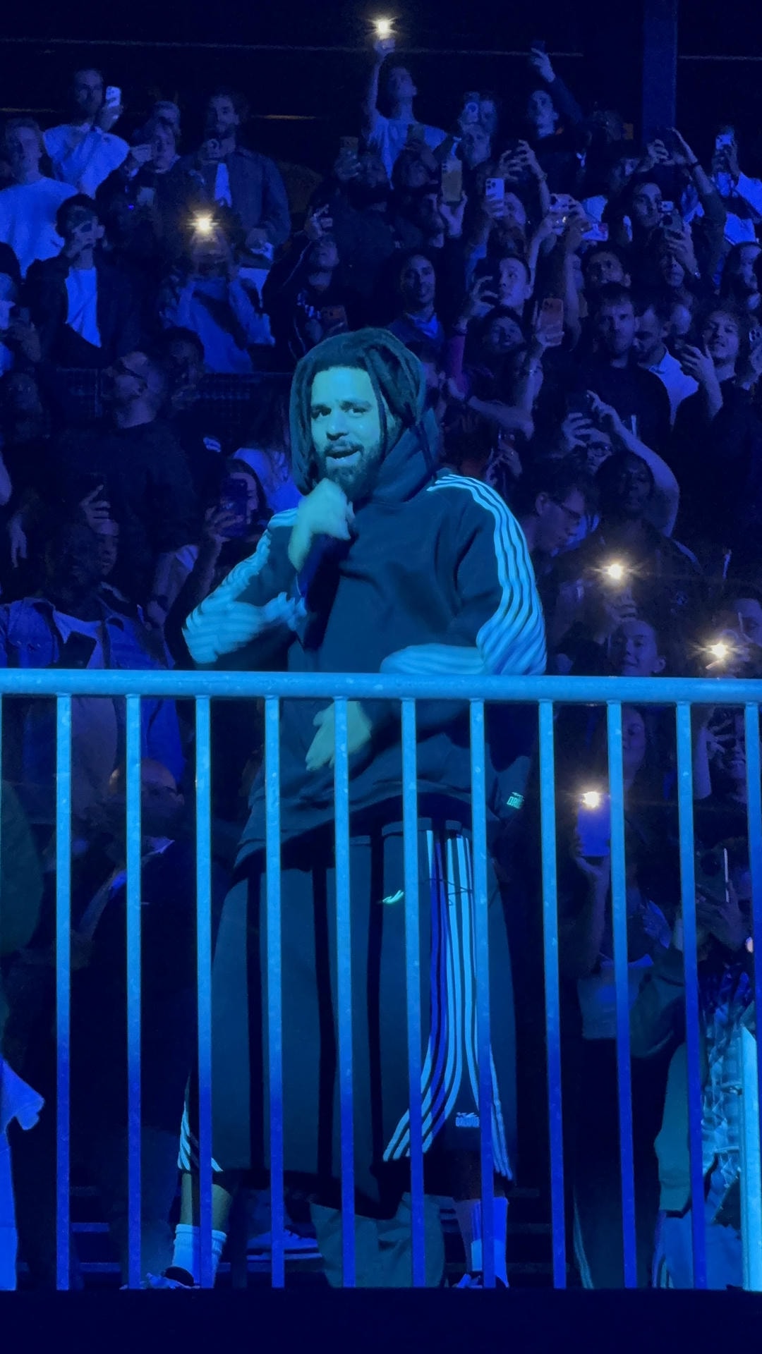 J. Cole standing in front of a crowd of seated people on a balcony, lit by blue spotlights performing with a mic in his hand. He's wearing a tracksuit and looking out into the camera.