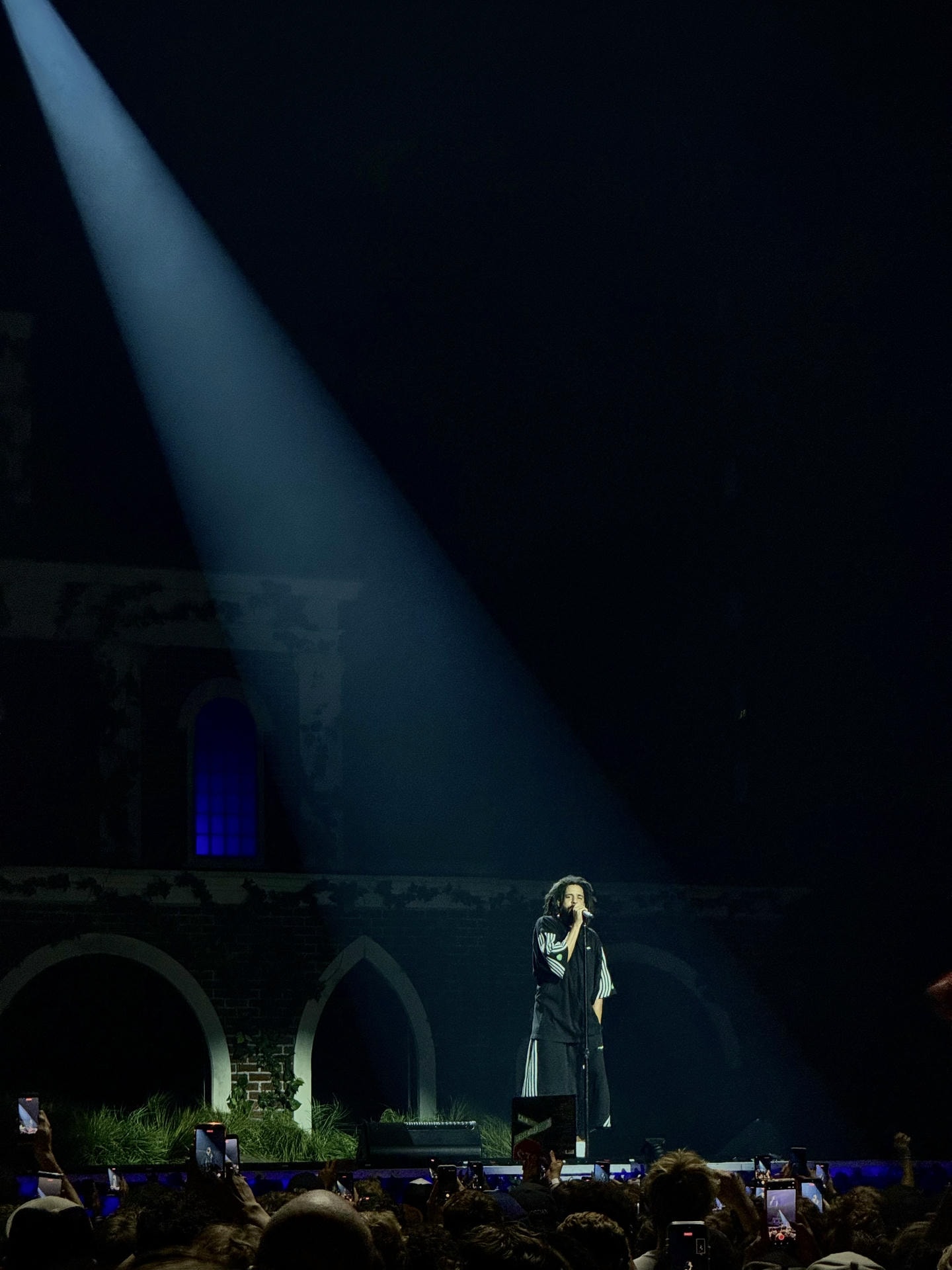 J. Cole performing on stage. He's lit by a spotlight from top left.